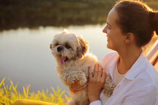 Retrato de perfil de una joven adulta sonriente de pelo oscuro positiva con camisa blanca y su perro pequinés sentado al atardecer cerca de las vacaciones en el río en la naturaleza