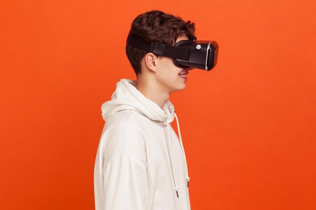 Retrato de perfil de un joven feliz con capucha de estilo informal disfrutando de la vista en gafas de realidad virtual, tecnologías modernas. Disparo de estudio interior aislado sobre fondo naranja