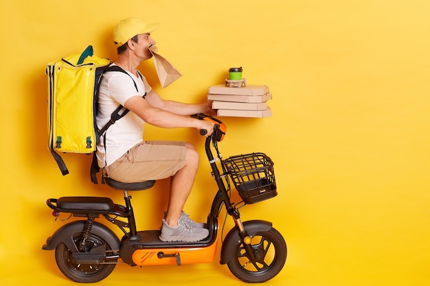 Retrato de perfil de un joven con mochila con camiseta blanca montando en scooter con cajas de pizza y café sosteniendo un paquete de papel en los dientes entregando muchos pedidos aislados sobre un fondo amarillo