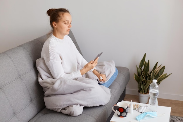Retrato de perfil de una mujer enferma sentada sobre la tos envuelta en una manta, usando el teléfono móvil para revisar los correos electrónicos o marcar el número de teléfono, sufrir influenza, resfriarse o tener gripe.