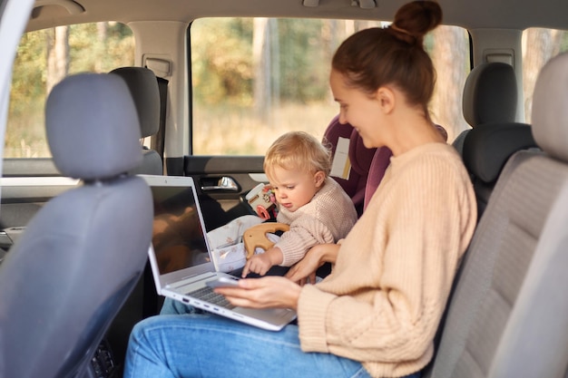 Retrato de perfil de una mujer feliz y positiva que trabaja en una laptop mientras se sienta y juega con su hija pequeña en una silla de seguridad en el asiento trasero del auto, un niño pequeño tratando de escribir en un cuaderno