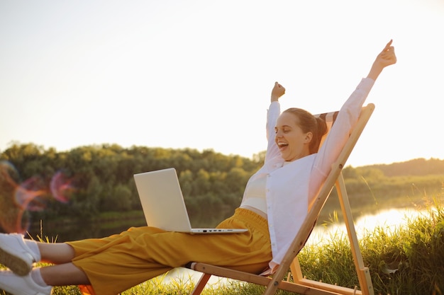 Retrato de perfil de una mujer feliz y satisfecha con camisa blanca sentada junto al agua en una silla plegable y trabajando en una laptop gritando felizmente celebrando su victoria y éxito