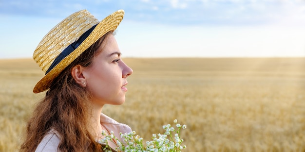 Retrato de perfil de una mujer joven con un sombrero de paja y con un ramo de flores silvestres en un campo de trigo al amanecer