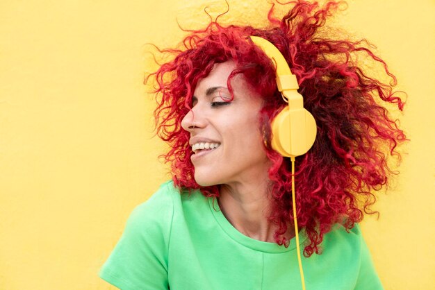 Foto retrato de perfil de una mujer latina sonriente con cabello afro rojo usando una camiseta escuchando música con auriculares amarillos sobre un fondo amarillo