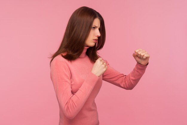 Retrato de perfil mujer morena concentrada sosteniendo los puños apretados, luchando con el abusador, defensa de la violencia doméstica. Disparo de estudio interior aislado sobre fondo rosa