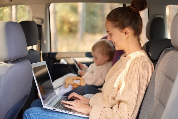 Retrato de perfil de una mujer sonriente y encantada que trabaja en una computadora portátil mientras se sienta con su hija en una silla de seguridad en el asiento trasero del automóvil