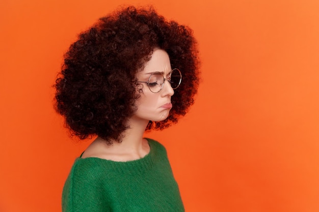 Retrato de perfil de una mujer triste y disgustada con peinado afro usando un suéter verde de estilo casual de pie con labios de puchero, siente tristeza, llora. Disparo de estudio interior aislado sobre fondo naranja.