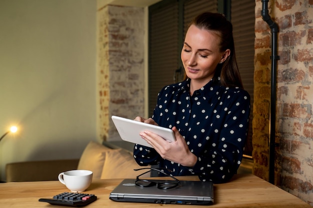 Retrato de un periodista alegre con ropa informal sentado en una mesa con un aparato en las manos usando Internet WiFi en una oficina moderna con interior