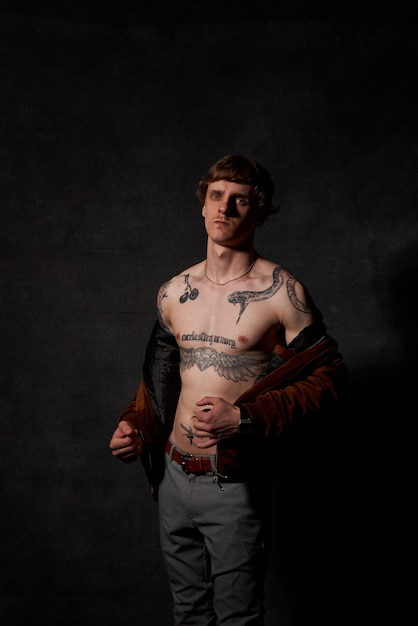 Retrato de una persona hipster con cuerpo tatuado en poses de chaqueta en fondo oscuro