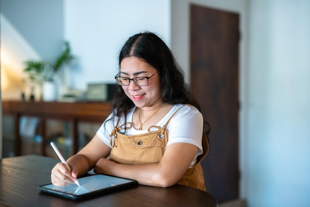 Retrato de personas independientes asiáticas mujeres de negocios con gafas elegantes hipste Dibuja o toma nota en una tableta digital con bolígrafo electrónico para navegar por Internet, chatear y bloguear en una cafetería