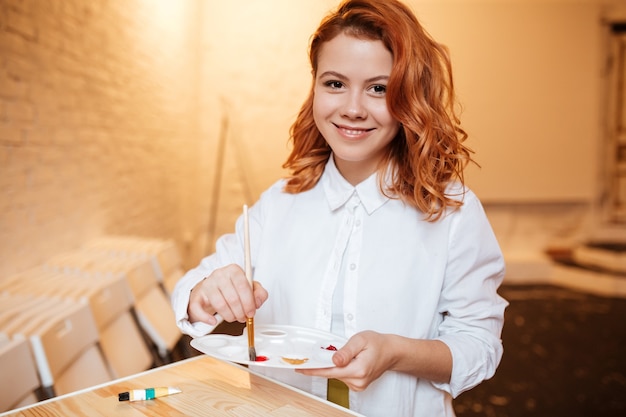 Retrato de pintor de mujer joven pelirroja feliz con pinturas al óleo y paleta.