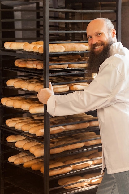 Foto retrato de placer joven panadero adulto con barba larga en uniforme blanco de pie cerca de los estantes llenos de galletas recién horneadas en su lugar de trabajo concepto de profesión interior mirando a la cámara