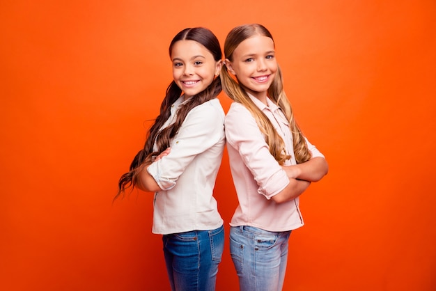 Retrato de positivo alegre equipo de dos niños lindos niñas stand back-to-back listo resolver soluciones escolares decidir decisiones llevar traje moderno aislado fondo de color naranja