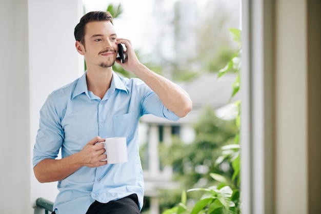 Retrato de positivo apuesto joven bebiendo café por la mañana y hablando por teléfono con un amigo