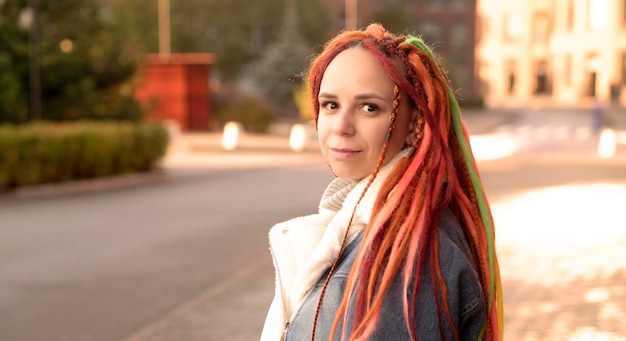 retrato, de, positivo, mujer joven, con, dreadlocks, en, moderno, calle de la ciudad, bastante, hembra, con, brillante, peinado, mirar lejos, sonriente