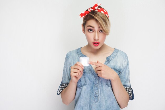 Retrato de preocupación hermosa mujer joven en camisa de mezclilla casual y diadema roja de pie, sosteniendo el condón y señalando con el dedo con cara de pregunta. Foto de estudio de interior, aislado sobre fondo blanco.