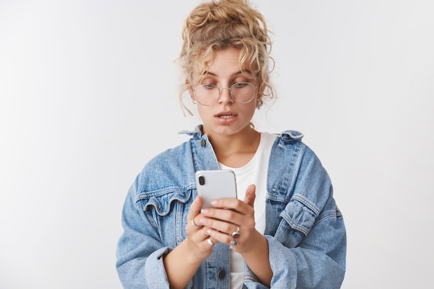 Retrato preocupado intenso bonito elegante moderno 20 anos jovem loira encaracolado bagunçado coque usar óculos jaqueta jeans parece perturbado nervoso tela do smartphone lendo mensagem, verificando a lista de compras