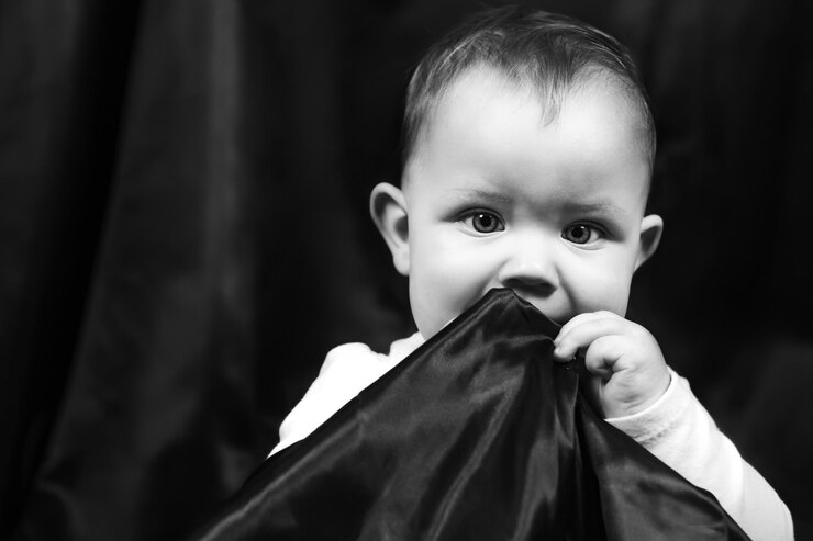 Foto retrato preto e branco de um bebê de um ano a criança se senta em um fundo de tecido preto coloca o tecido de fundo na boca com uma mão olha para a câmera com grandes olhos atônitos