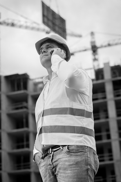 Retrato preto e branco do jovem engenheiro de construção, falando por telefone no canteiro de obras. guindaste levantando blocos pesados no fundo