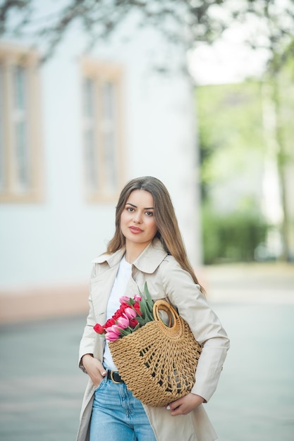 Retrato de primavera de una joven hermosa mujer feliz de 28 años con el pelo largo y bien peinado sostiene una bolsa de mimbre en sus manos con un ramo de tulipanes en una calle de la ciudad Modelo elegante en una gabardina