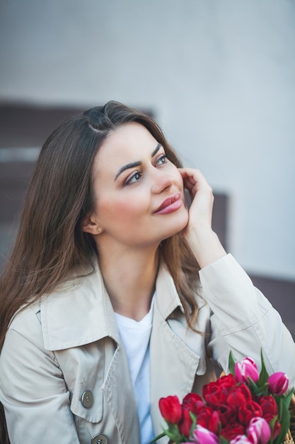 Retrato de primavera de una joven hermosa mujer feliz de 28 años con el pelo largo y bien peinado sostiene una bolsa de mimbre en sus manos con un ramo de tulipanes en una calle de la ciudad Modelo elegante en una gabardina