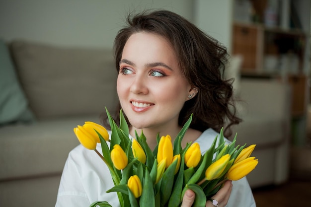 Retrato de primavera de una joven hermosa mujer con ojos azules y cabello rizado con ramo de tulipanes amarillos en el interior de una casa Modelo feliz en jeans y camisa blanca Día de la Madre