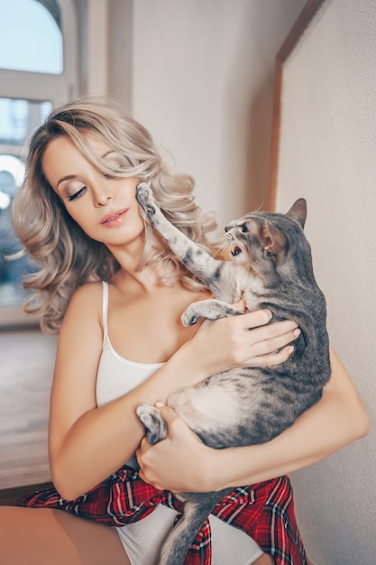 Retrato del primer del dueño feliz joven hermoso hermoso de la mujer rubia y del gato agresivo adorable de la lucha divertida en el interior de la casa. Peterbald pelo corto oriental gris sphynx. Concepto de personas y animales