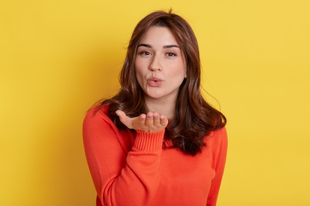 El retrato del primer de la hembra encantadora atractiva de aspecto agradable, la muchacha alegre coqueta hace pucheros los labios que envían beso suave del aire sobre amarillo, mira la cámara.