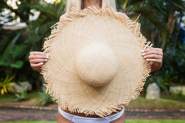 Retrato del primer de la mujer joven que sostiene el sombrero de paja grande en la playa tropical