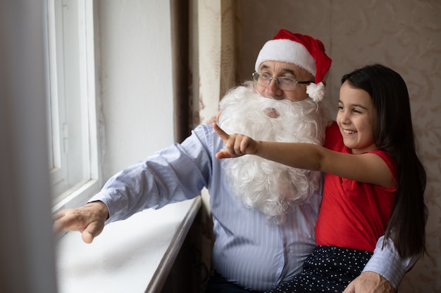 Retrato de primer plano de abuelo abrazos niña alegre en sombreros. abuelo y nieta con sombreros de Santa Claus