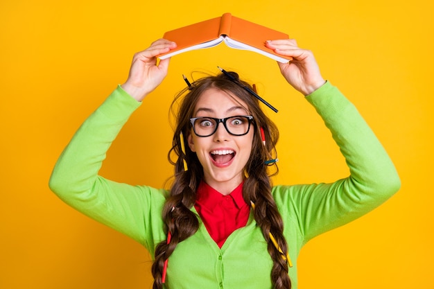 Foto retrato de primer plano de una adolescente alegre bastante funky sosteniendo un libro de ejercicios sobre la cabeza como divertirse aislado sobre fondo de color amarillo brillante