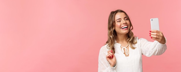 Retrato de primer plano de una alegre y optimista chica rubia sonriente con un vestido blanco riéndose como un amigo grabando videollamadas en una aplicación móvil tomando una foto selfie en el fondo rosa del teléfono inteligente