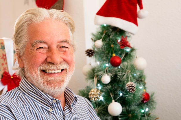 Retrato y primer plano del anciano maduro sonriendo y riendo mirando a la cámara con un árbol de Navidad en el fondo - ancianos jubilados en el día de Navidad en casa