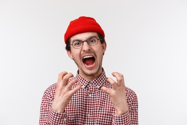 Retrato de primer plano de angustiado y enojado joven inconformista gracioso con bigote llevar gorro rojo, gafas, apretar las manos molestas y molestas, gritando de enojo,