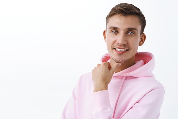 Retrato de primer plano de un apuesto y creativo cabello rubio masculino y una sudadera con capucha rosa que sostienen la mano cerca de la barbilla y sonríen complacidos tienen una excelente idea de pie pensativo como una sugerencia interesante mira la cámara