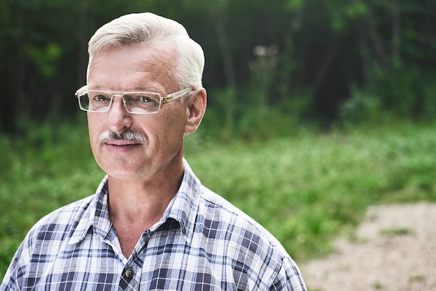 Retrato de primer plano de un apuesto hombre adulto canoso con bigote y gafas sonriendo en el fondo de un parque forestal de verano