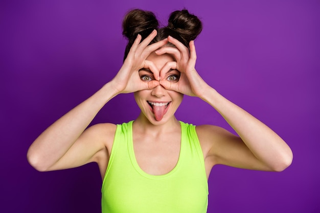 Retrato de primer plano de la atractiva chica alegre tonta que muestra el signo de ok como gafas con la lengua fuera aislado sobre fondo de color púrpura violeta brillante