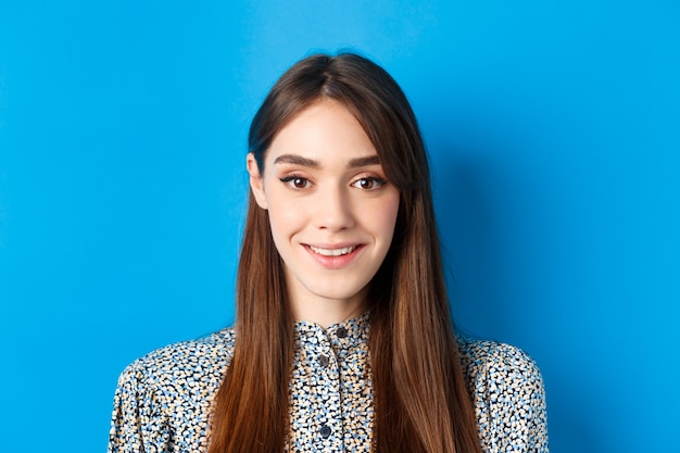 Retrato de primer plano de una atractiva chica natural con cabello largo y maquillaje, sonriendo feliz y amigable a la cámara, azul.