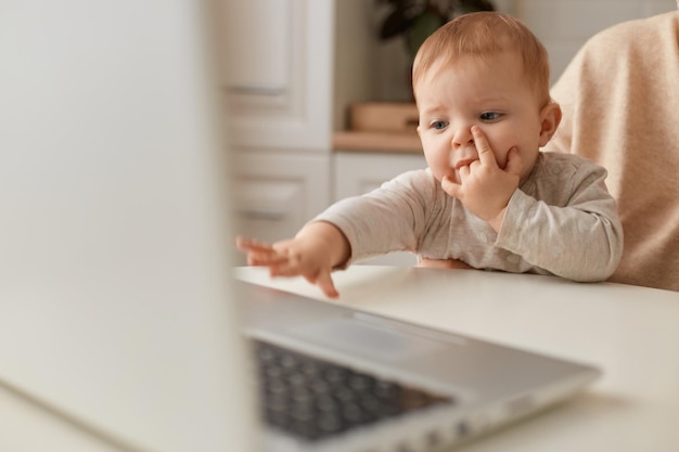 Retrato de primer plano de un bebé sentado en las rodillas de la madre sin rostro y extendiendo su mano a los niños de la computadora portátil y la tecnología moderna