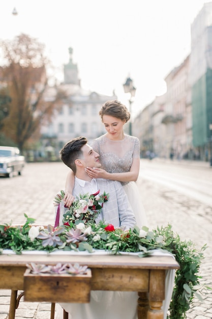 Retrato de primer plano de la bella novia y el novio guapo, abrazándose y sentado en la mesa decorada, fondo de la ciudad vieja