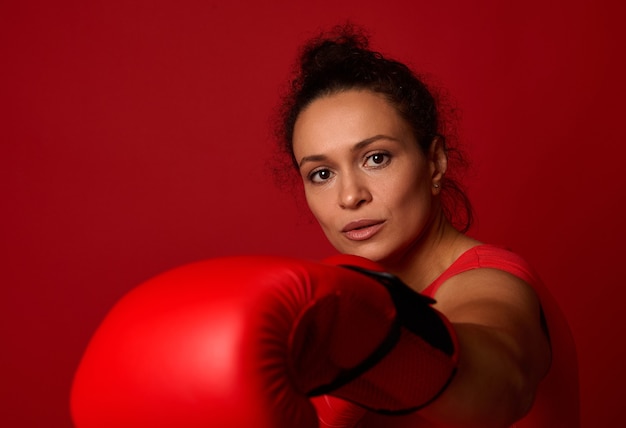 Retrato de primer plano de la boxeadora joven concentrada de deportes con guantes de boxeo rojos, haciendo un golpe directo, golpeando hacia la cámara, aislado sobre fondo rojo con espacio de copia. Concepto de arte marcial