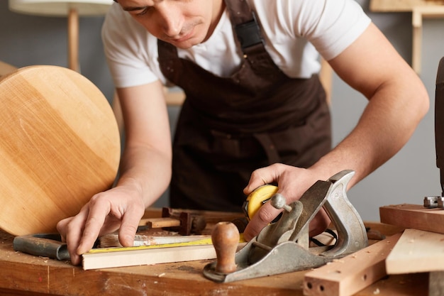Retrato de primer plano de carpintero irreconocible con delantal y camiseta blanca que mide el tablón posando entre diferentes equipos de carpintería que trabajan solo en el taller