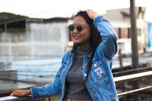 Retrato de primer plano de una chica de cabello largo que se ríe con jeans de chaqueta y gafas de sol en el fondo de la ciudad Chica inteligente de pie en el puente Concepto de estilo de vida