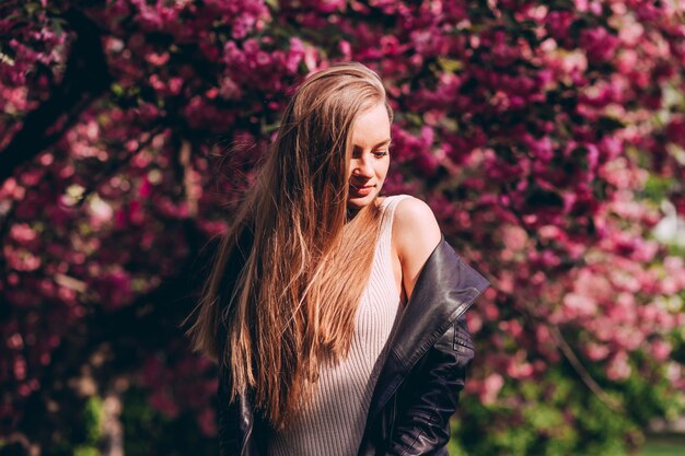 Un retrato de primer plano de una chica rubia con el telón de fondo de un árbol de sakura japonés en flor. Una hermosa joven con largo cabello rubio en el parque de la primavera.