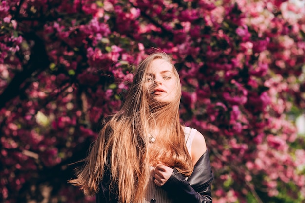 Un retrato de primer plano de una chica rubia con el telón de fondo de un árbol de sakura japonés en flor. Una hermosa joven con largo cabello rubio en el parque de la primavera.