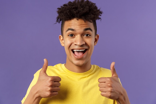 Retrato de primer plano de un chico inconformista animado y entusiasta con corte de pelo afro que está de acuerdo con algo, muestra el pulgar hacia arriba sonriendo con alegría, recomienda un buen producto, le gusta y aprueba el plan