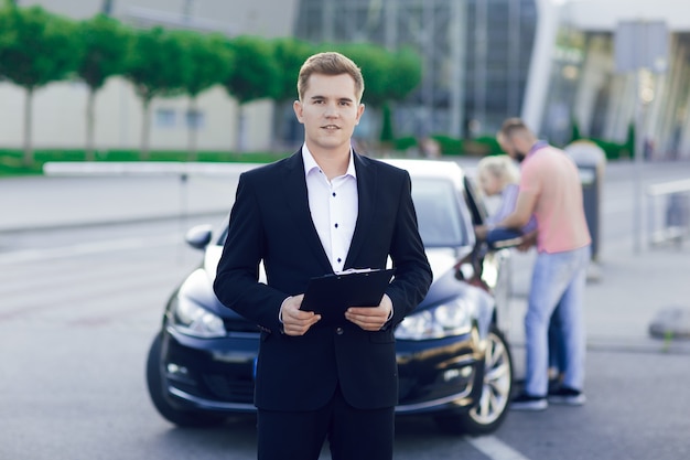 Retrato de primer plano de un concesionario de automóviles joven en un traje de negocios. Detrás de él, una pareja joven, un hombre y una mujer inspeccionan un coche nuevo. Compra de máquinas, prueba de manejo.