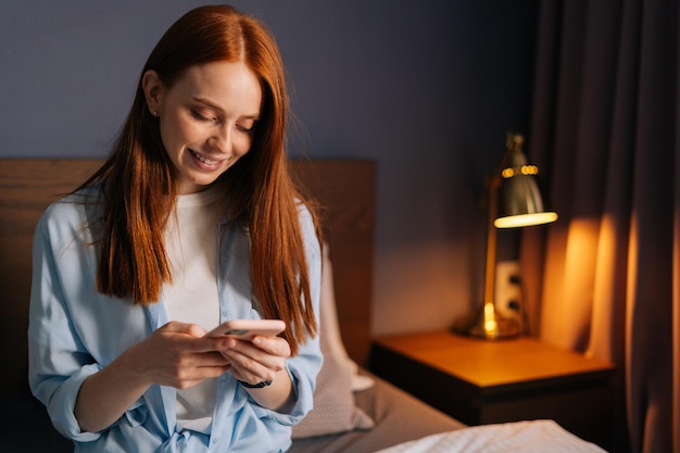Retrato de primer plano de una encantadora joven pelirroja escribiendo un mensaje en el teléfono móvil en el apartamento