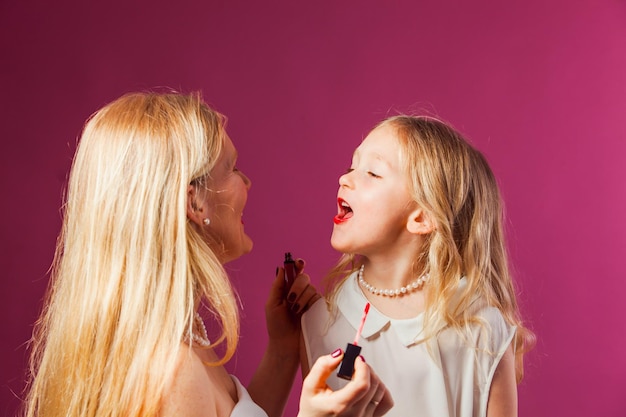 Retrato de primer plano de encantadora madre e hija rubias en estudio Mujer coloreando labios de niñas con lápiz labial rojo líquido brillante Niña preescolar quiere ser como su madre