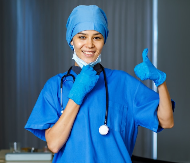 Retrato de primer plano de feliz médico de libertad bastante femenina en traje de hospital azul con estetoscopio sonriendo pulgar hacia arriba cuando se quita la mascarilla quirúrgica usada mira a la cámara mientras termina la pandemia.